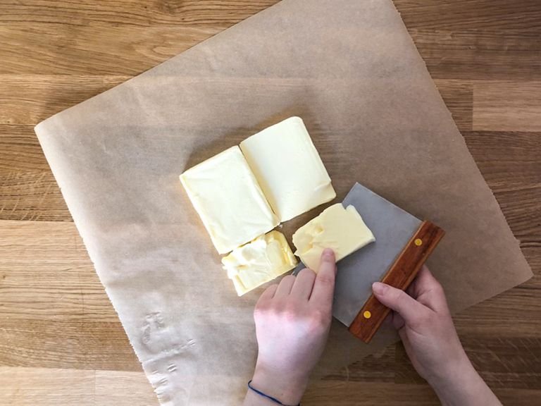 Nun kann die Butter vorbereitet werden. Einen Bogen Backpapier auslegen, Butter in vier Teile schneiden und als Quadrat darauflegen. Einen weiteren Bogen Backpapier darüber legen, dann die Butter mit dem Nudelholz ausrollen, so dass ein Quadrat von ca. 16x16 cm entsteht. Mit dem Backpapier einschlagen und über Nacht im Kühlschrank lagern.
