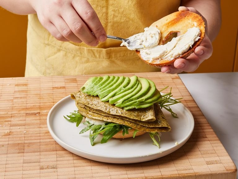 To build the sandwich, add arugula to the base of the bagel, then shave Parmesan on top. Place the omelette on top of that, then fan out the sliced avocado over the eggs. Spread the lid of the bagel with the lemony cream cheese and place on top. Serve and enjoy!