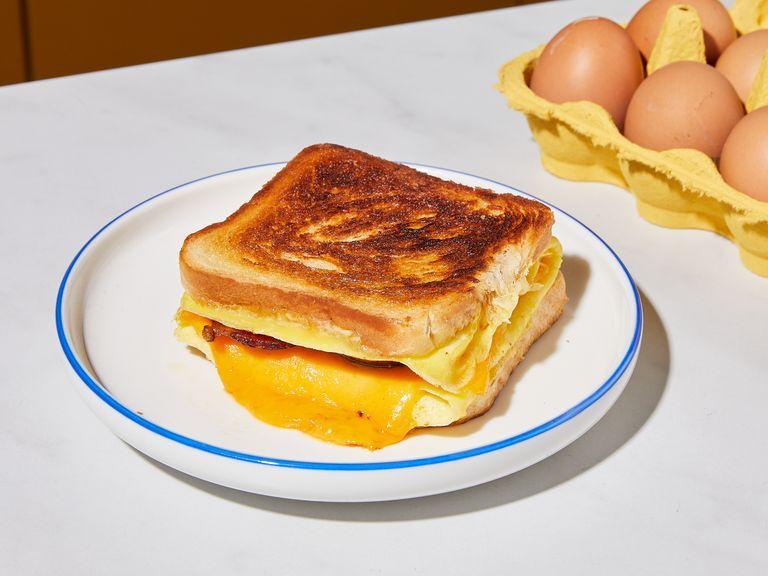 To make the one-pan egg sandwich, place bacon in a cold skillet, then fry over medium heat until crispy on both sides. Remove and drain on paper towels. Whisk eggs with a pinch of salt in a bowl. Butter one side of each of the two slices of toast. Set skillet over low heat with remaining bacon grease and add whisked eggs. Swirl pan until egg is completely and evenly distributed, then place slices of toast, buttered-side up, in the center of the egg so that the "bottoms" are facing each other, but leaving approx. 2 cm/1 in. of space between them (watch video for visual instruction). Fry until the egg is completely set, then flip the whole thing over with a spatula. Fold the egg on all sides onto the two slices of toast, place bacon and cheese on top and fry for approx. 2 min. more. Fold the sandwich and fry again on both sides for approx. 2 min., or until golden brown. Enjoy immediately!