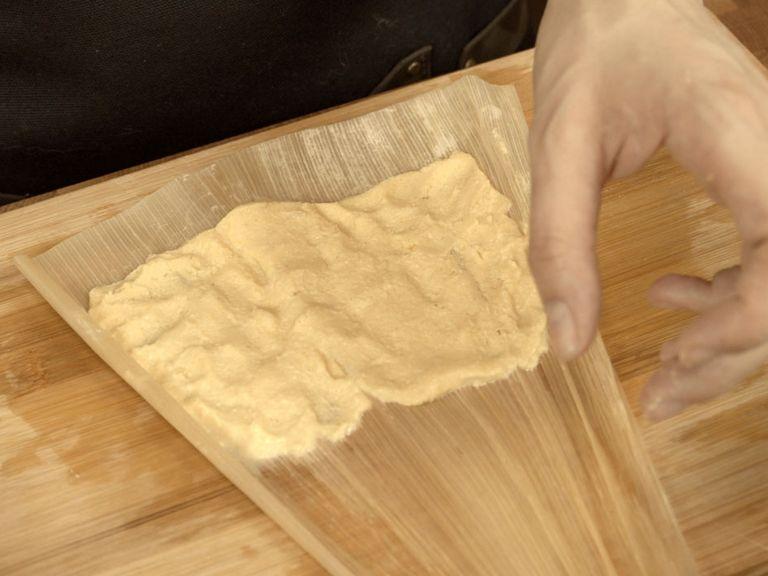 Start assembling the tamales. Lay corn a soaked corn husk glossy side up. Add 2 tbsp of masa dough in the wider half of the corn husk. Spread it evenly in the wider half, leave 1-2 cm gap from top and sides. Tip: dip your fingers in a bowl with clean water, it will prevent dough from sticking