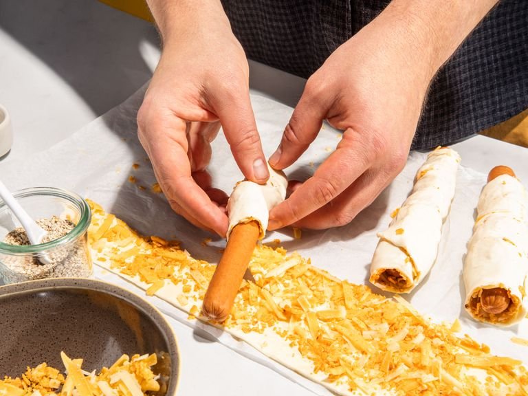 Blätterteig ausrollen und mit dem verquirlten Ei bestreichen. Gleichmäßig mit der Tortilla Chips-Käse Mischung bestreuen und leicht andrücken. Dabei etwas der Mischung als Topping zurückbehalten. Blätterteig in 6 lange Streifen schneiden und jeweils ein Hot-Dog-Würstchen in einem Streifen Blätterteig einrollen. Die eingerollten Hot-Dog-Würstchen jeweils mit einem scharfen Messer in 4 gleiche Stücke teilen.