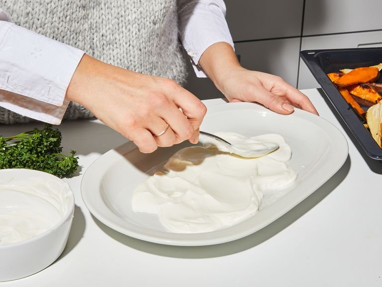 Das Gemüse aus dem Ofen nehmen. Den Knoblauch entnehmen und das cremige Innere vorsichtig in eine Schüssel pressen. Mit einer Gabel zu einer Paste zerdrücken. Die Knoblauchpaste mit einem Schneebesen in die warme Haselnuss-Butter-Soße einrühren und gut vermischen. Zum Anrichten den griechischen Joghurt auf einer großen Servierplatte verteilen. Darauf das geröstete Wurzelgemüse geben und mit der braunen Haselnussbutter beträufeln. Mit Zitronenschale garnieren, krause Petersilie darüber zupfen und mit flockigem Meersalz bestreuen. Guten Appetit!