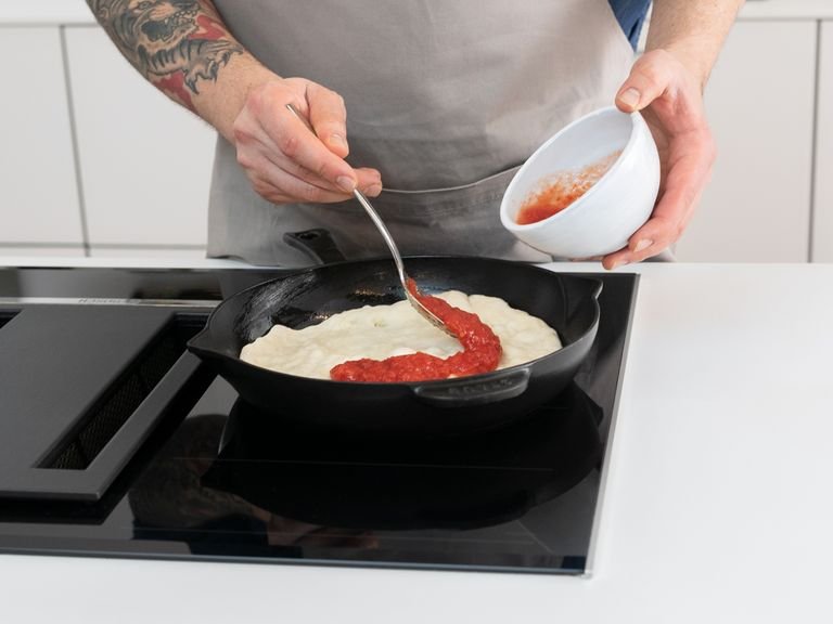 Sobald der Teig gegangen ist, den Ofen auf 250°C vorheizen und das Grillrost auf oberster Schiene in den Ofen schieben. Den Teig auf einer leicht bemehlten Arbeitsfläche leicht durchkneten, bis er ungefähr die Größe der gusseisernen Pfanne hat, in der die Pizza gebacken wird. Jetzt die Gusseisenpfanne auf mittlerer bis hoher Stufe erhitzen. Sobald die Pfanne heiß ist, das restliche Olivenöl hinzufügen und den Teig in die Pfanne geben. Alles für ca. 1 Min. anbraten, dann die Tomatensoße mit einem Löffel gleichmäßig auf dem Teig verteilen.