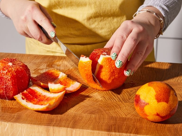 In der Zwischenzeit die Schale von 2 Blutorangen abreiben und die Schale in eine große Schüssel geben. Dann alle Blutorangen schälen. Dafür mit einem großen Messer die Ober- und Unterseite abschneiden, sodass die Orange flach auf dem Brett sitzen kann. Dann im Kreis herum die Schale und das Weiß vom Fruchtfleisch abschneiden. Die Orangen vierteln und die Viertel wieder halbieren, sodass du mundgerechte Stücke erhältst. Zur Seite stellen. Rote Zwiebel in feine Ringe schneiden.