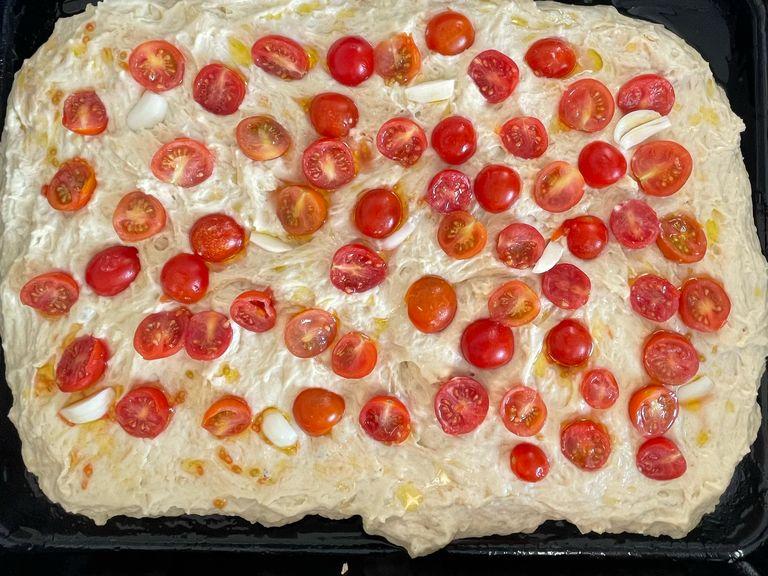 Once the resting time is over, we can place our focaccia in a baking tray. Cover the tray with a table spoon of oil, then lay the dough in it and spread it with your hands. Add the sliced tomatoes, the sliced garlic and cover with another table spoon of oil (you can also add some coarse salt on top).