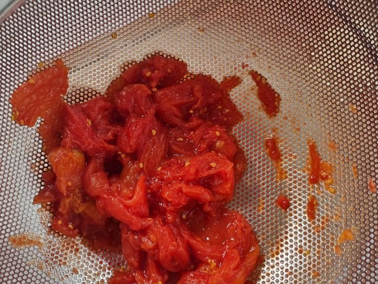 In einem großen Topf Wasser zum Kochen bringen und salzen. Knoblauch mit dem Messer zerdrücken. Dosentomaten in ein Sieb geben und die Flüssigkeit auffangen. Die Tomaten dabei mit den Fingern leicht zerdrücken.