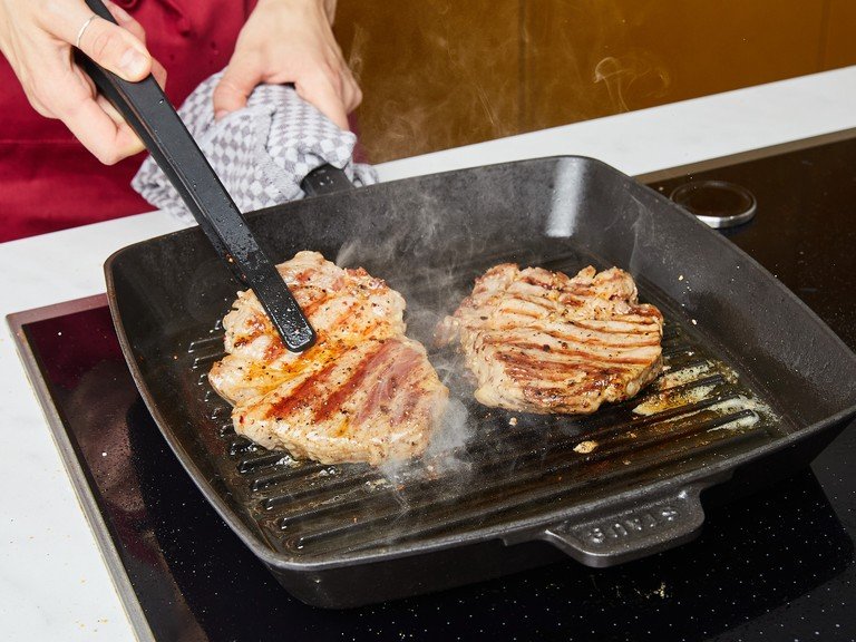 Sobald der Grill heiß ist, die marinierten Steaks mit dem restlichen Olivenöl bestreichen. Die Steaks auf den Grill legen. Achtung, während das Schweinefleisch gart, wird das Fett schmelzen, was zu leichtem Aufflammen führen kann. Die Steaks häufig bewegen, um ein Anbrennen zu vermeiden. Nach ca. 4 Min. Grillzeit die Steaks wenden und weitere ca. 3 Min. grillen, bis beide Seiten eine schöne Farbe angenommen haben. Auf ein Schneidebrett legen und ca. 5 Min. ruhen lassen.